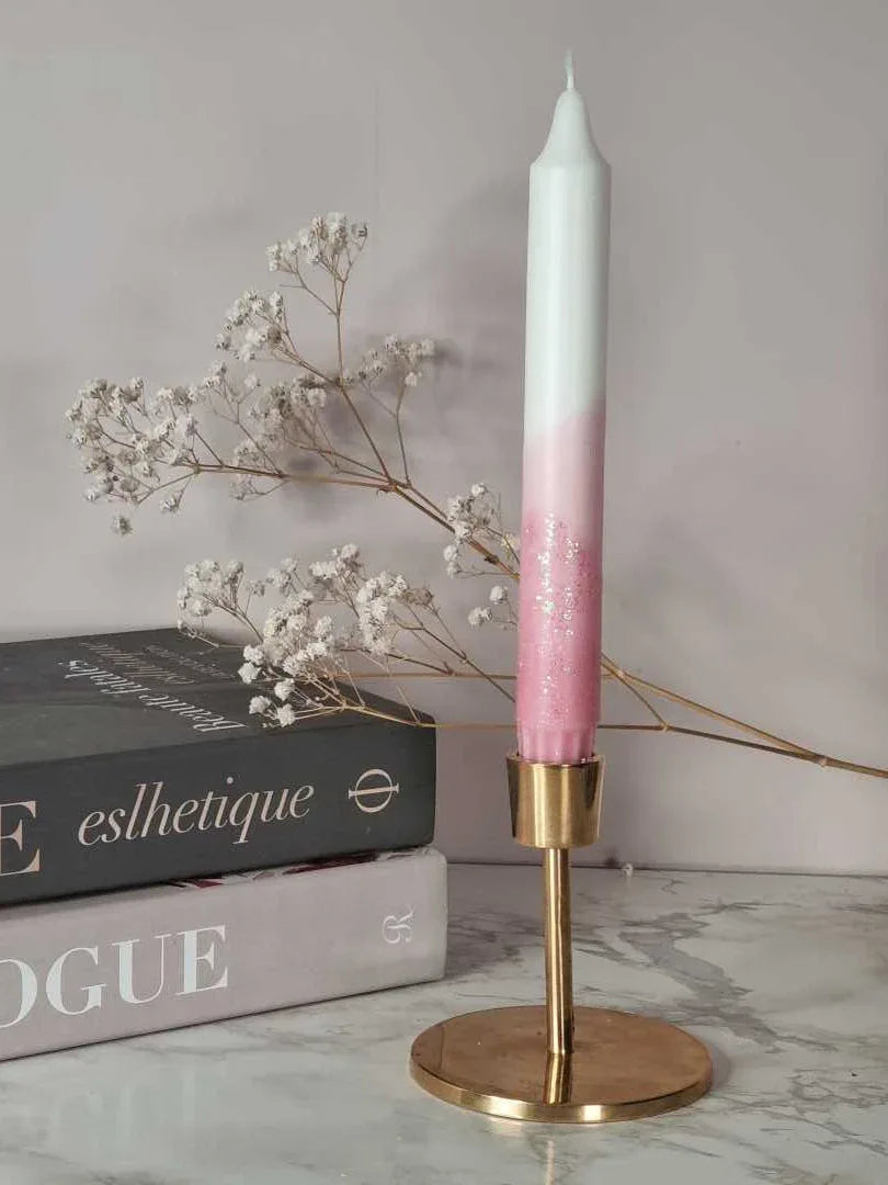 Dipdye kronelys i fargen rosa og hvitt med glitter. Lyset står i en messingstake. I bakgrunnen er det bøker og brudeslør